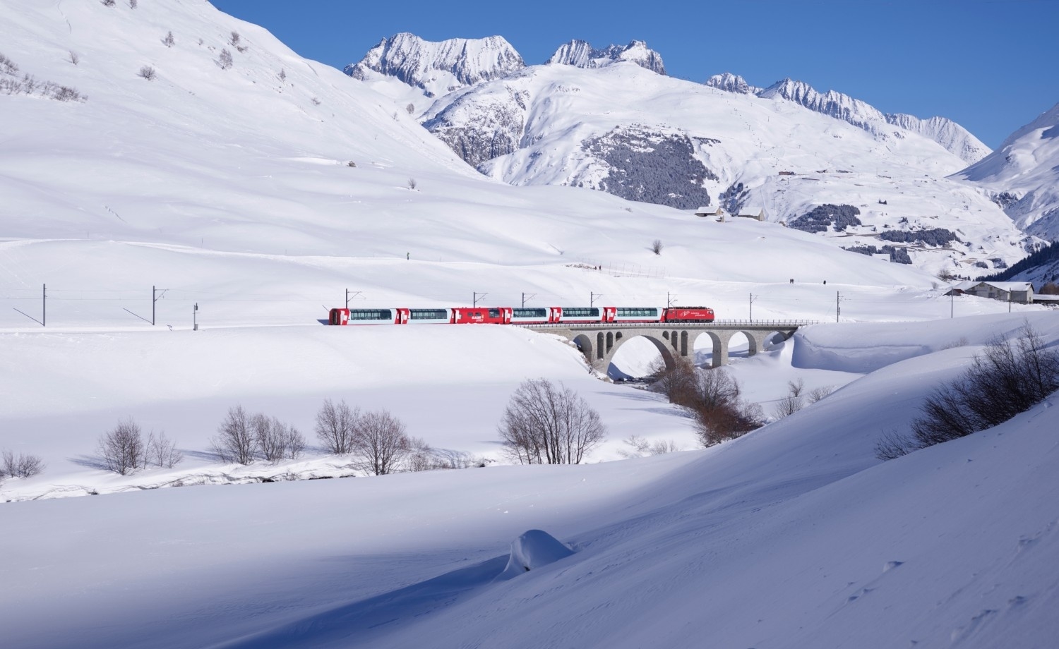 The image shows the Glacier Express train crossing a snow-covered valley with a stone bridge and mountain backdrop.