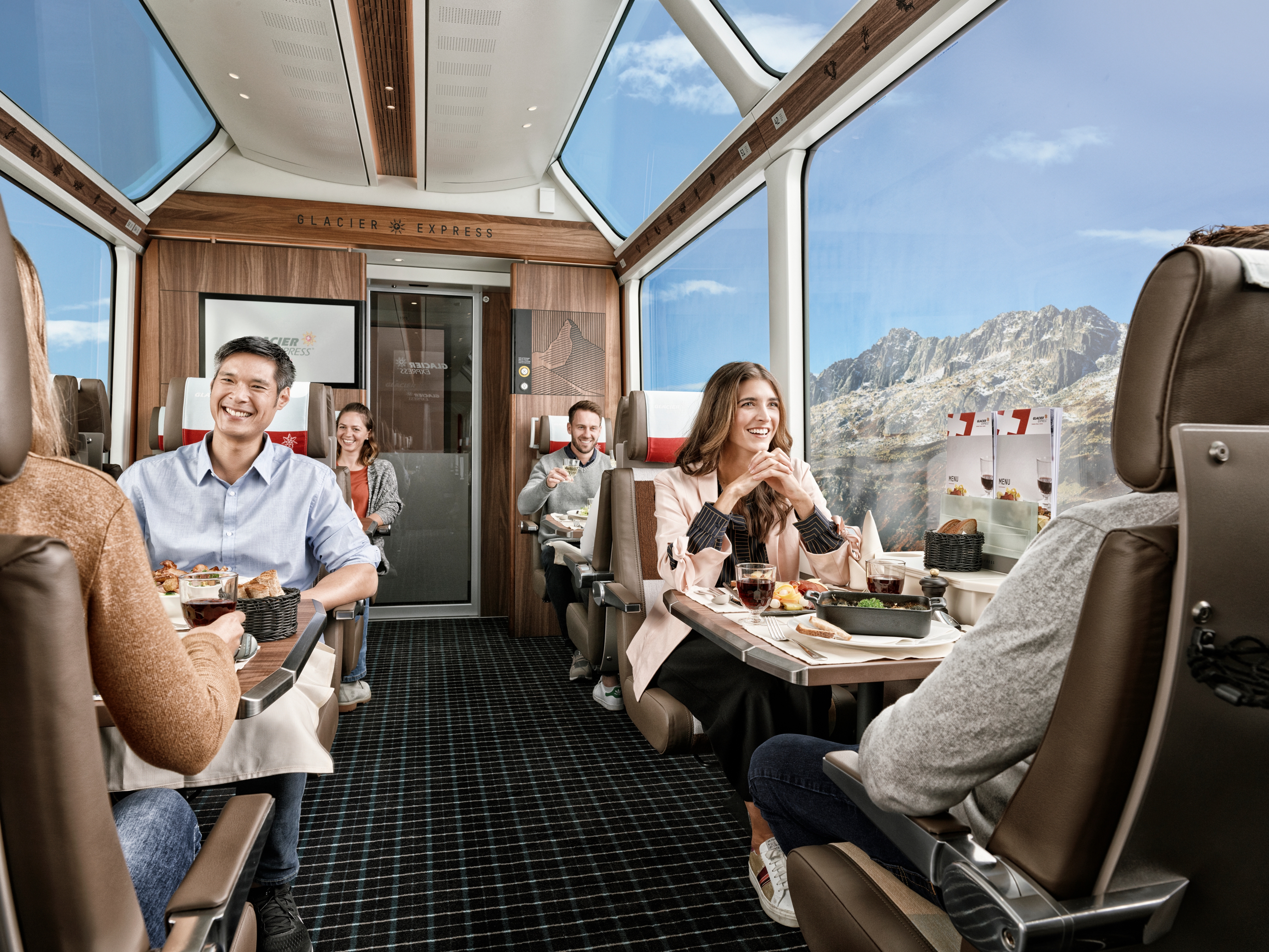 The picture shows the Glacier Express train where passengers are enjoying a meal with mountain views through panoramic windows.