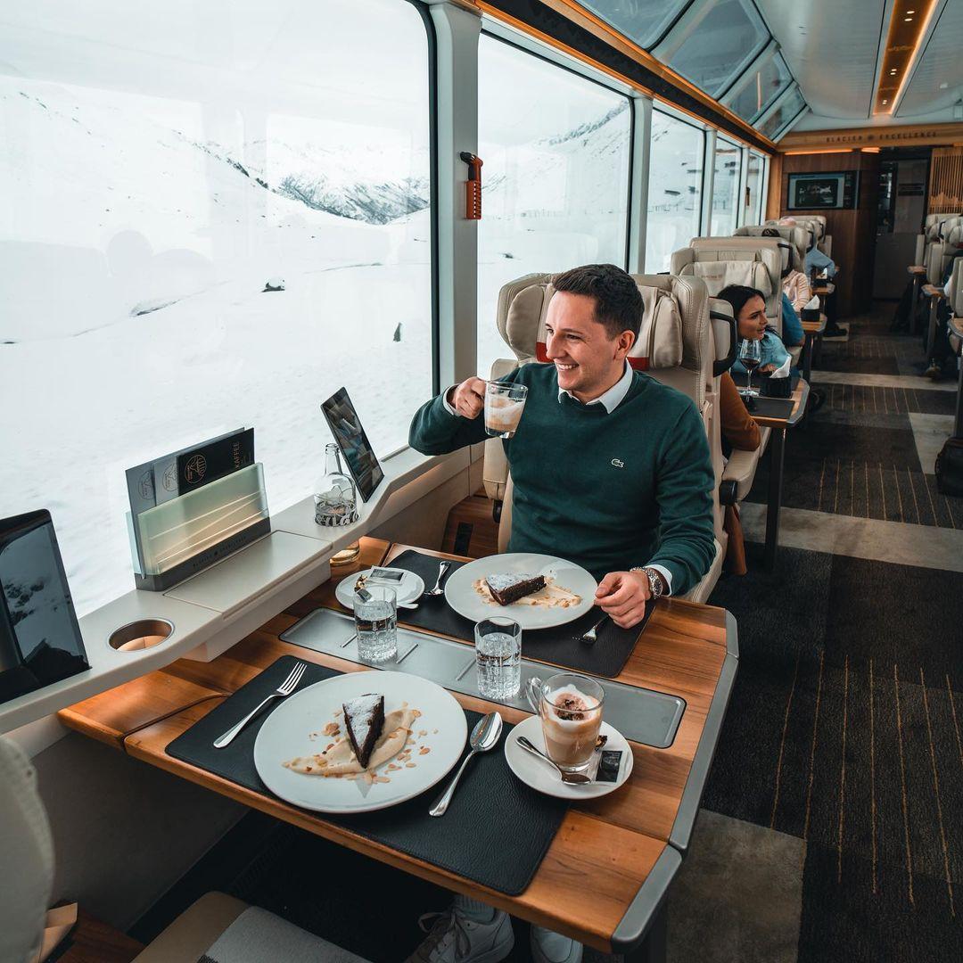 The image shows a passenger on the Glacier Express train drinking coffee with a dessert.