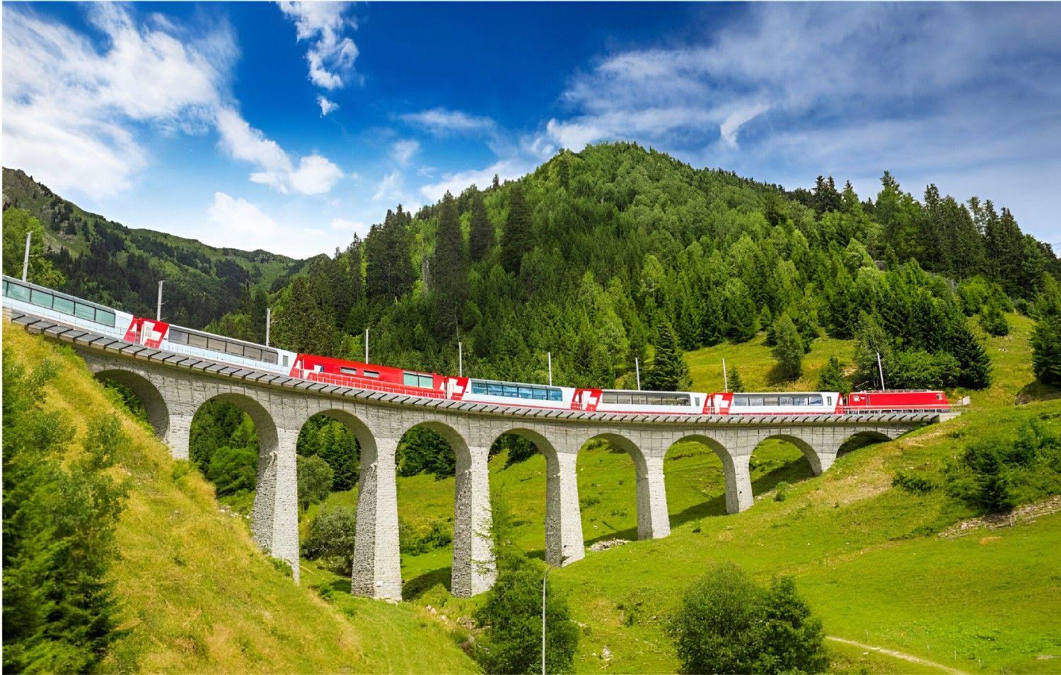 Auf dem Bild ist der Glacier Express zu sehen, der über eine Steinbrücke vor einer grünen Berglandschaft und blauem Himmel fährt.