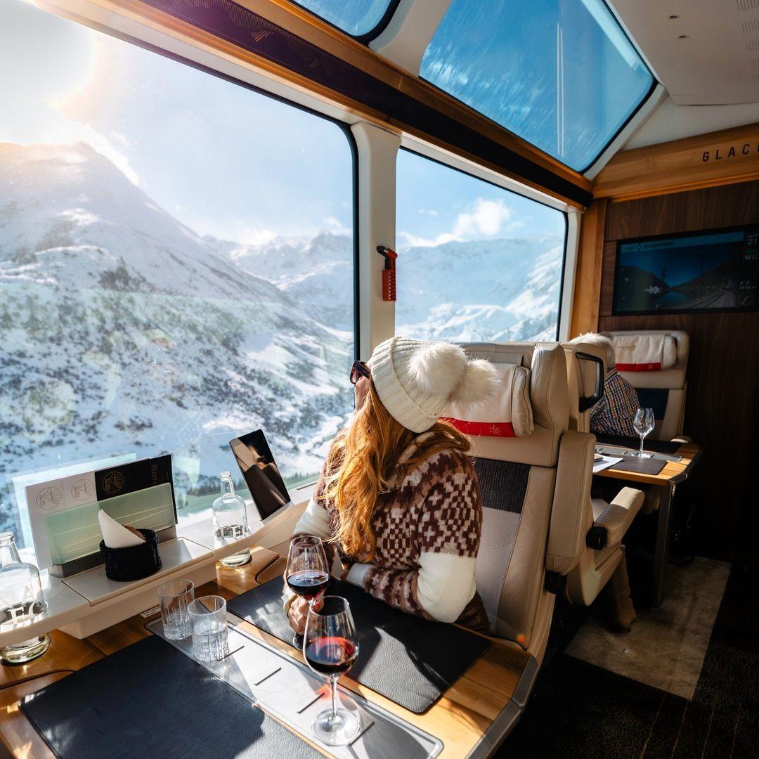 The image shows a girl passenger on the Glacier Express train looking at snow-capped mountains with a glass of wine.