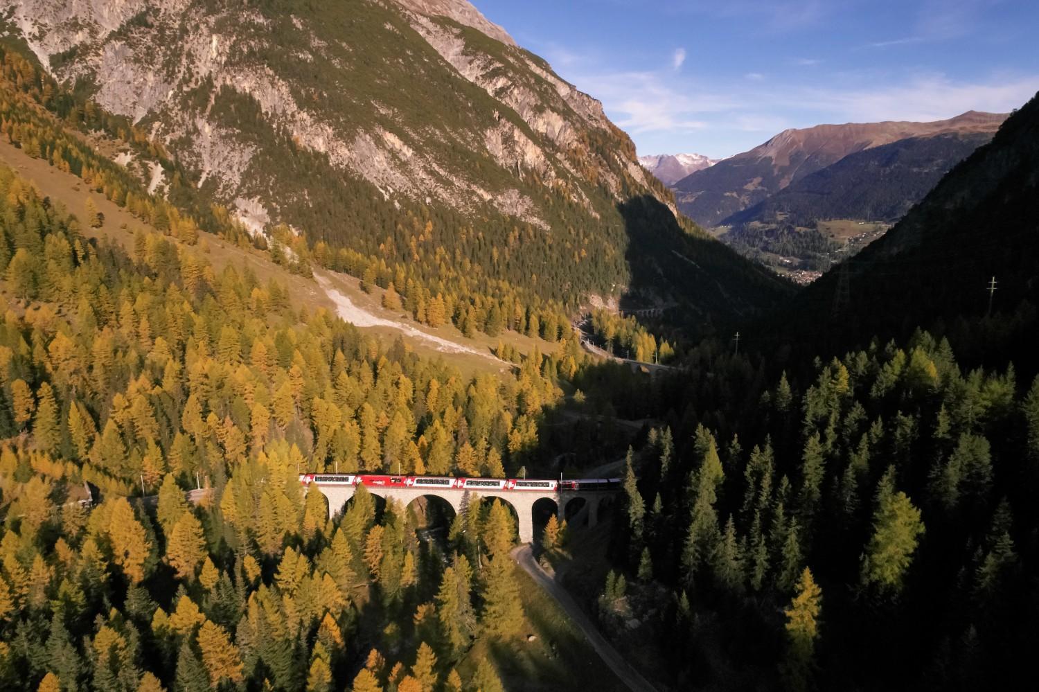 L'image montre le train Glacier Express sur un pont en pierre entouré d'arbres d'automne et de montagnes sous un ciel clair.