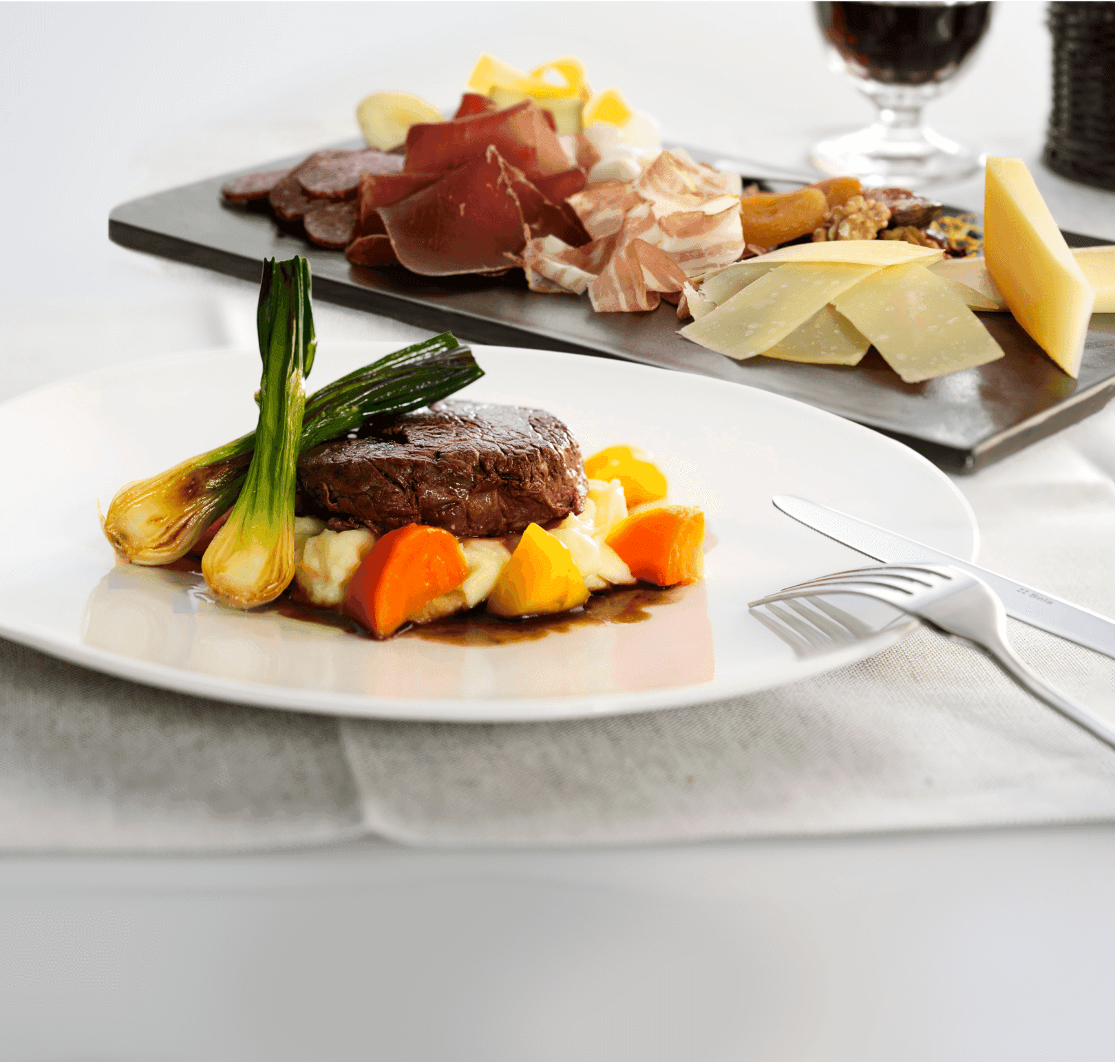 The image shows a gourmet steak with mashed potatoes and roasted vegetables. A charcuterie board with cheeses and meats is in the background.