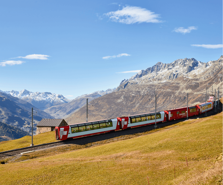 The picture shows the Glacier Express train traveling through a mountain landscape under a clear blue sky.