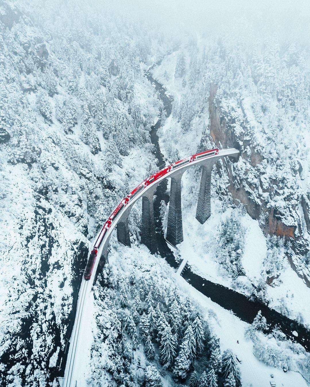 The image shows the Glacier Express train traveling over a stone bridge amidst snow-capped mountains.