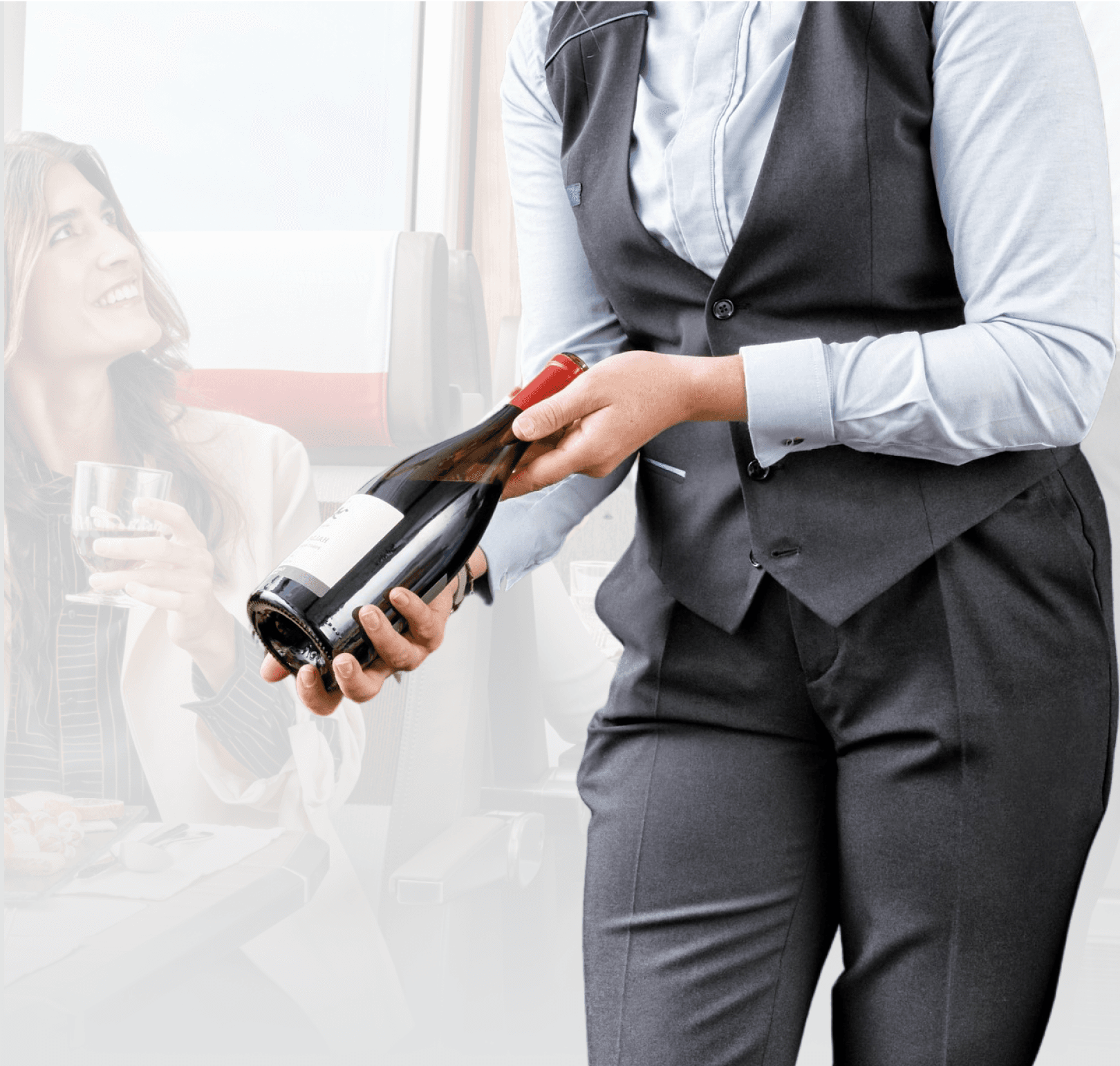 The image depicts a cheerful scene inside a train, where a server in a stylish uniform is pouring wine for a smiling female passenger. The environment suggests a comfortable and sociable train journey, enhancing the travel experience with attentive service and a pleasant atmosphere.