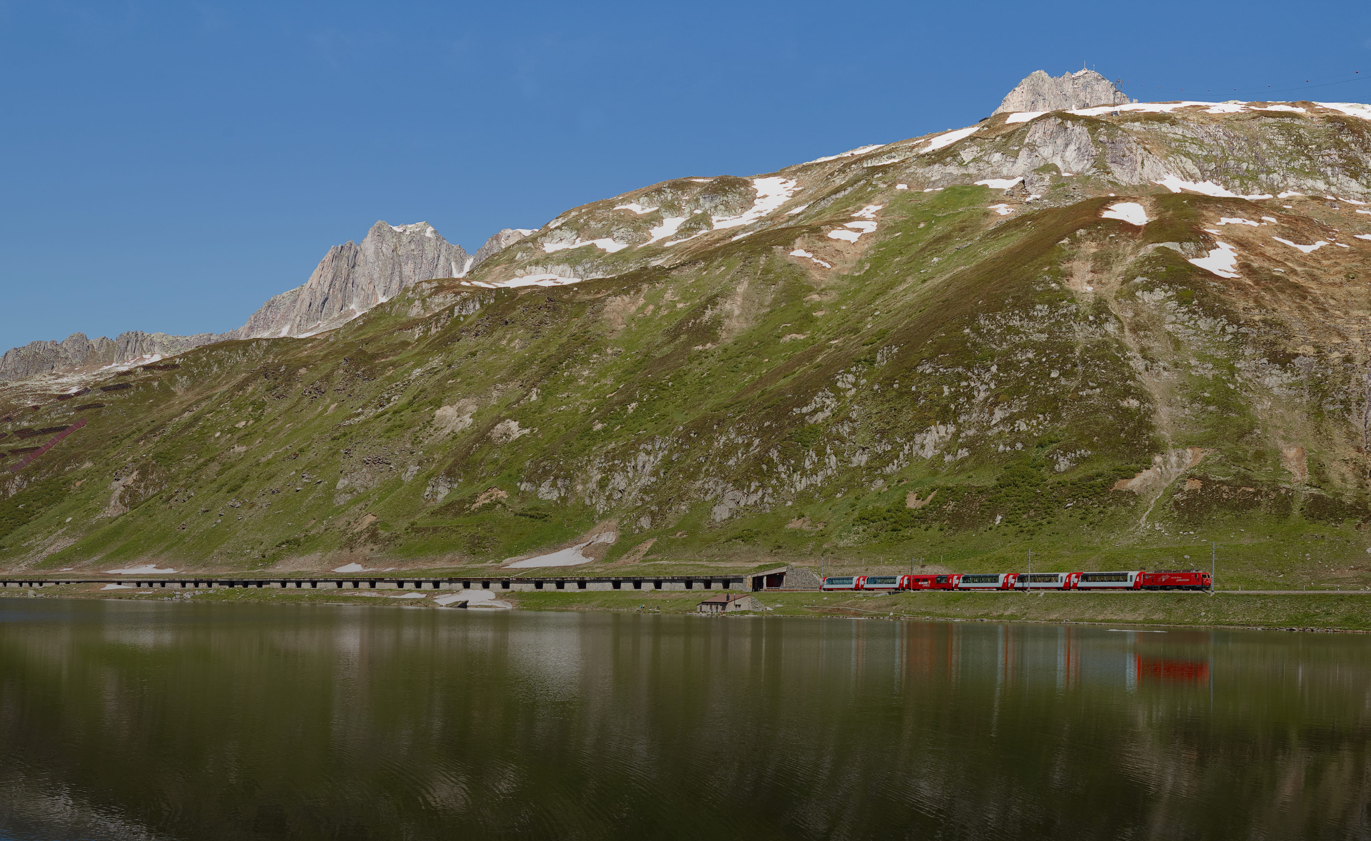  L'image montre le train Glacier Express longeant un lac avec des montagnes et des sommets rocheux sous un ciel clair.